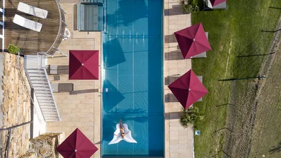 Vogelperspektive eines Pools mit einer Person auf einer Luftmatratze und roten Sonnenschirmen