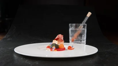 Kunstvoll angerichtetes Gourmetgericht auf weißem Teller mit Glas und Reagenzglas im Hintergrund