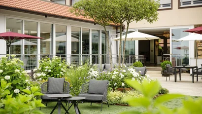 Gemütliche Terrasse mit Gartenmöbeln und blühenden Pflanzen vor Gebäude mit großen Fenstern.