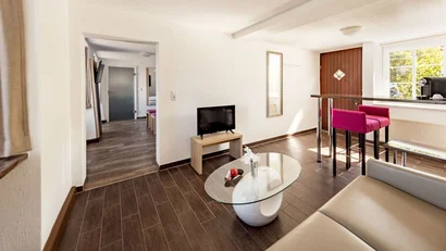 Heller Wohnraum mit Ledersofa, Glastisch, Fernseher und hohem Tisch mit pinken Stühlen