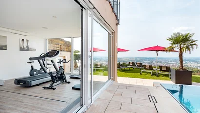 Fitnessraum mit Laufband und Fahrrad neben Terrasse mit Pool und Liegestühlen